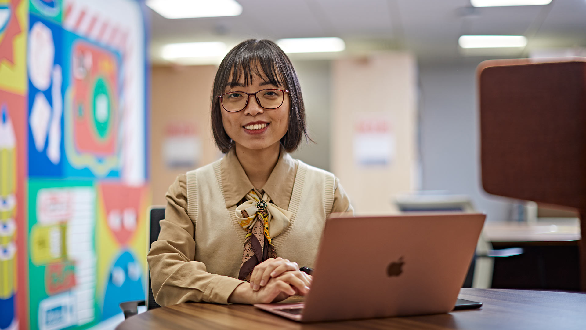 A student sat at a table in a library, with a laptop device.