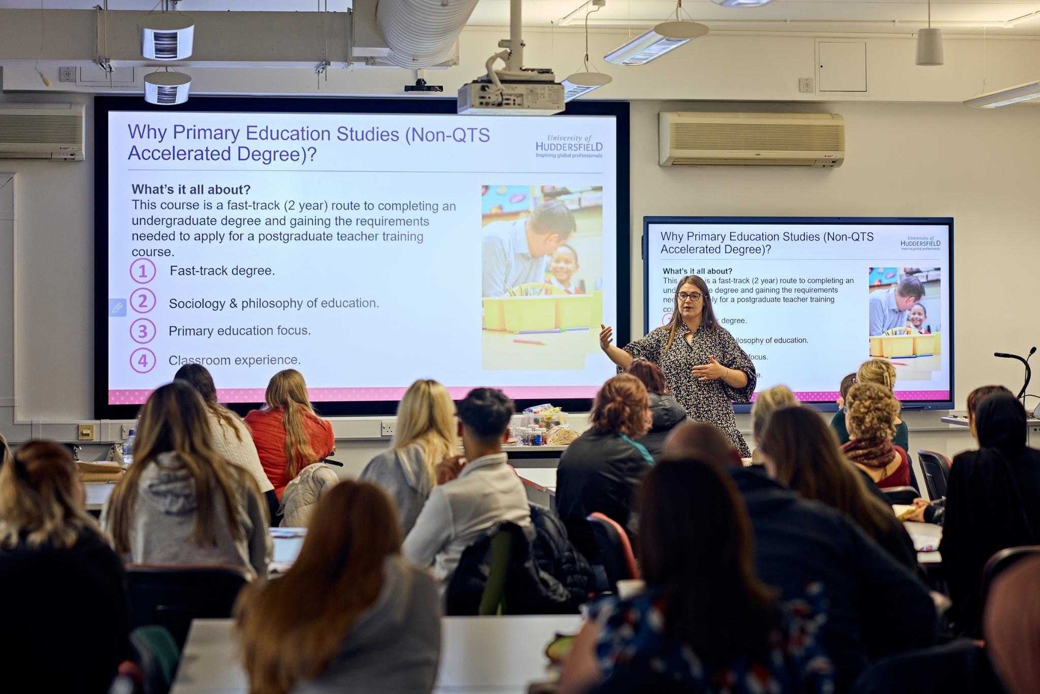 Female teacher presenting a lesson to a room of students, stood in front of two large screens