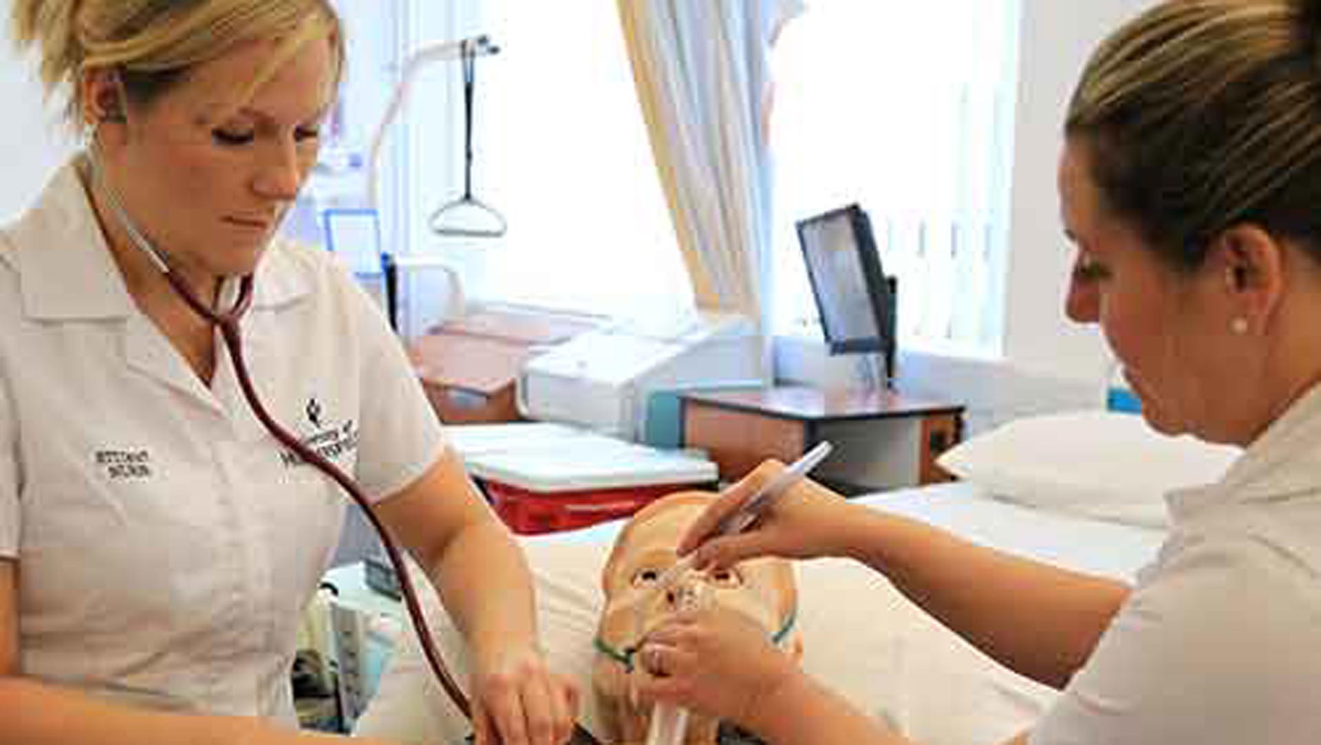 Adult nursing ward with a simulation dummy being treated on by 2 students.