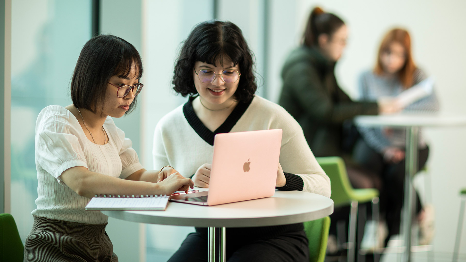 Two students sat at a table working on a laptop device.