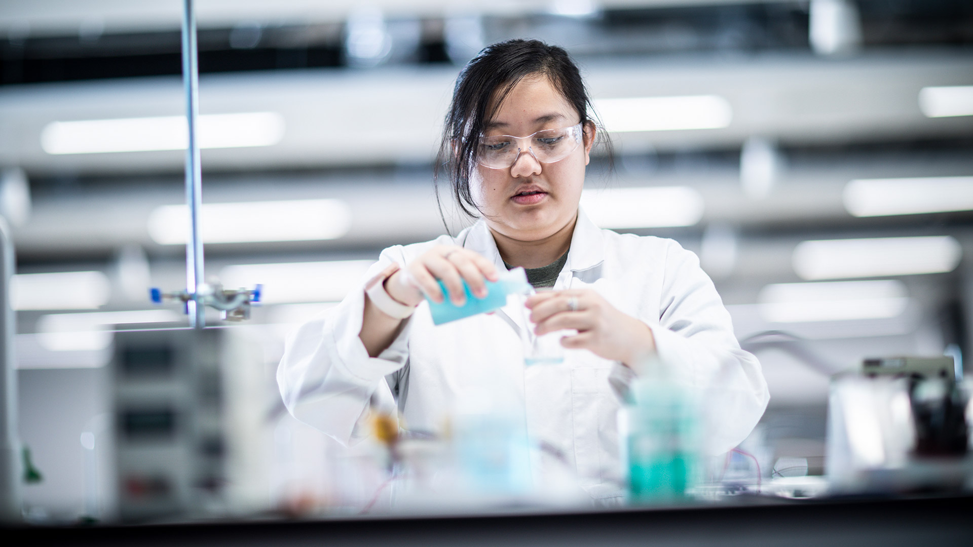 A student working with chemicals in a lab. They are wearing a lab coat and glasses.