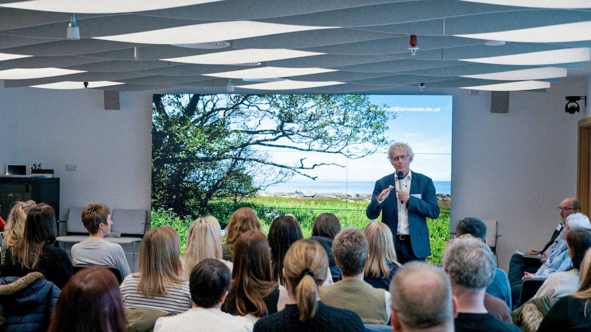 A man in a suite talks to a room full of people. Behind him is a presentation showing a tree and the countryside.