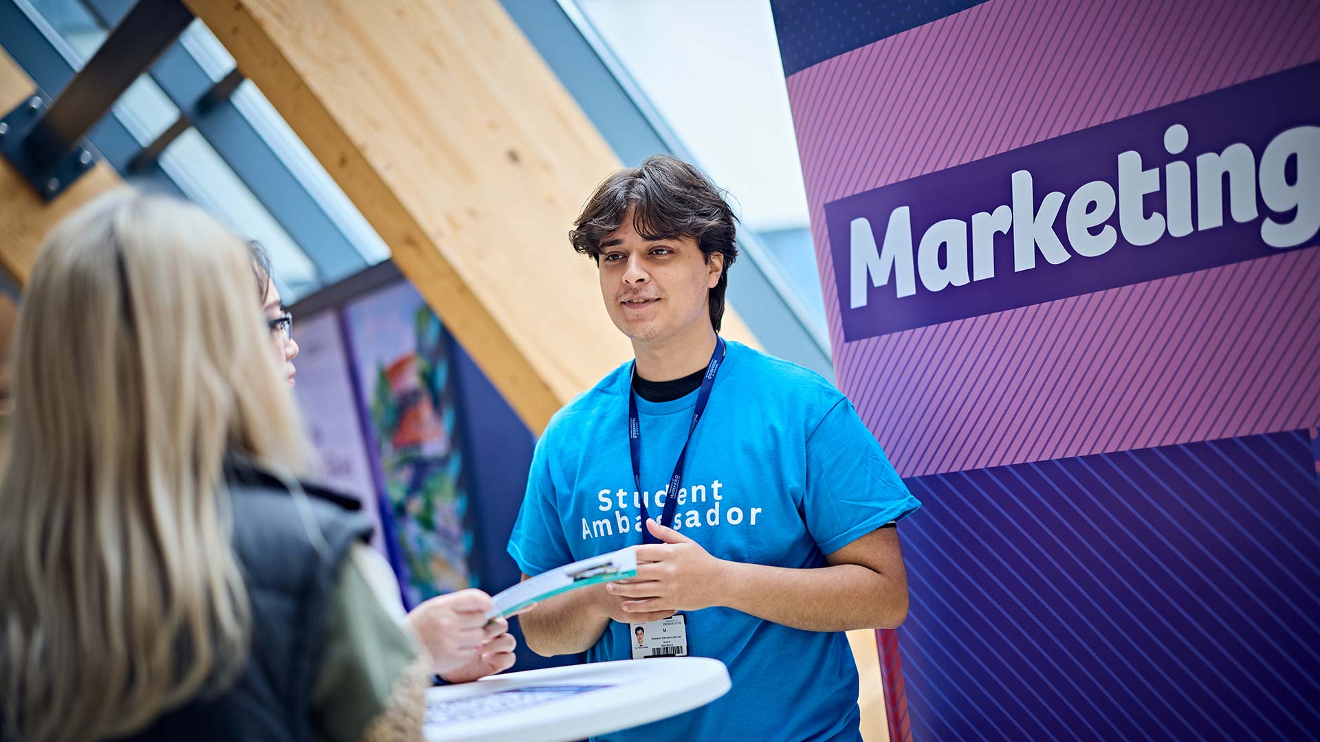 Mohammad talking to a guest at an Open Day. There is a colourful pull-up banner behind him that says 